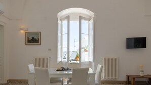 A dining area with a white table and chairs, a painting on the wall, a large window with a view, and a TV mounted on the wall.