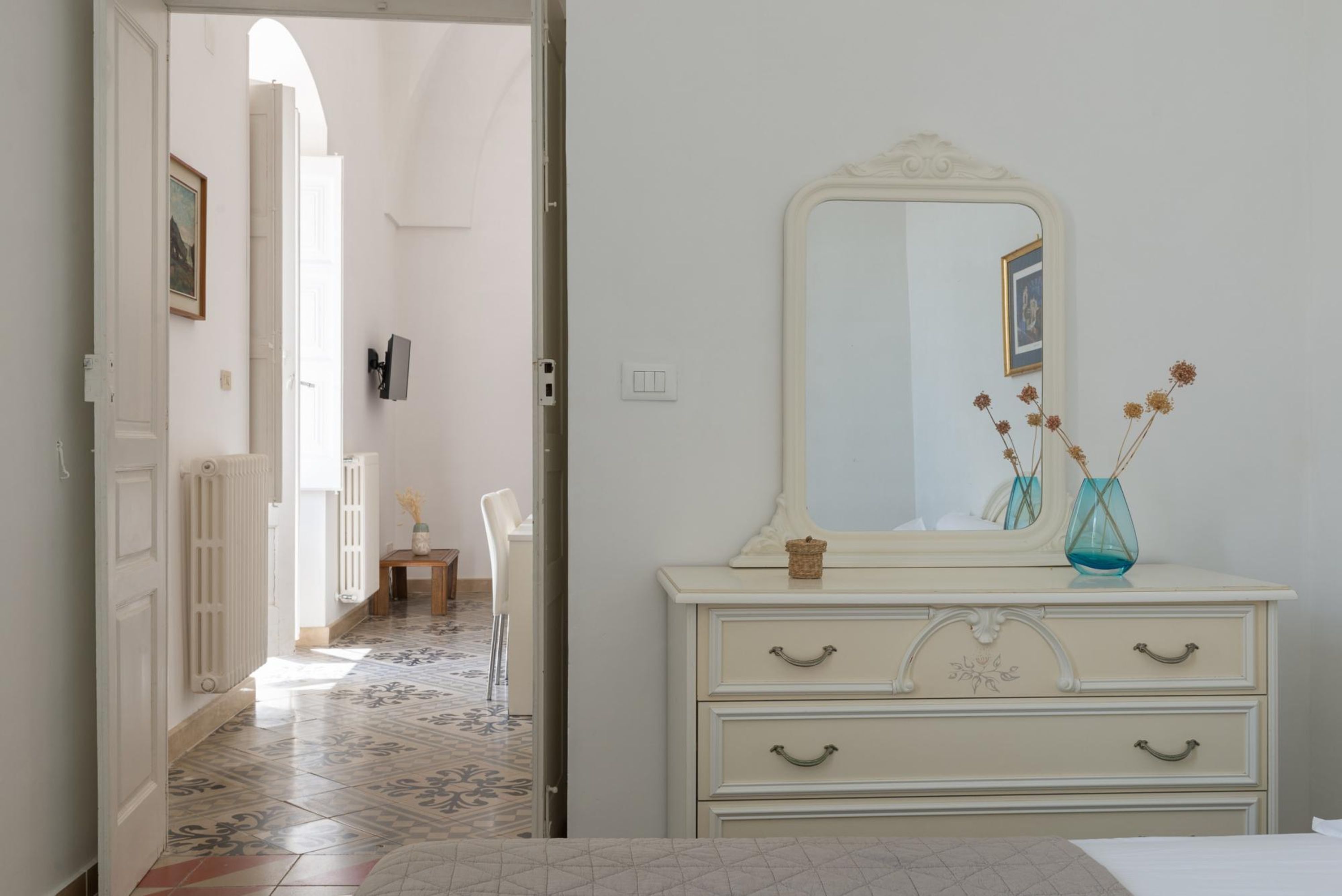 Een kamer met een witte dressoir, een spiegel en een vaas met gedroogde bloemen.