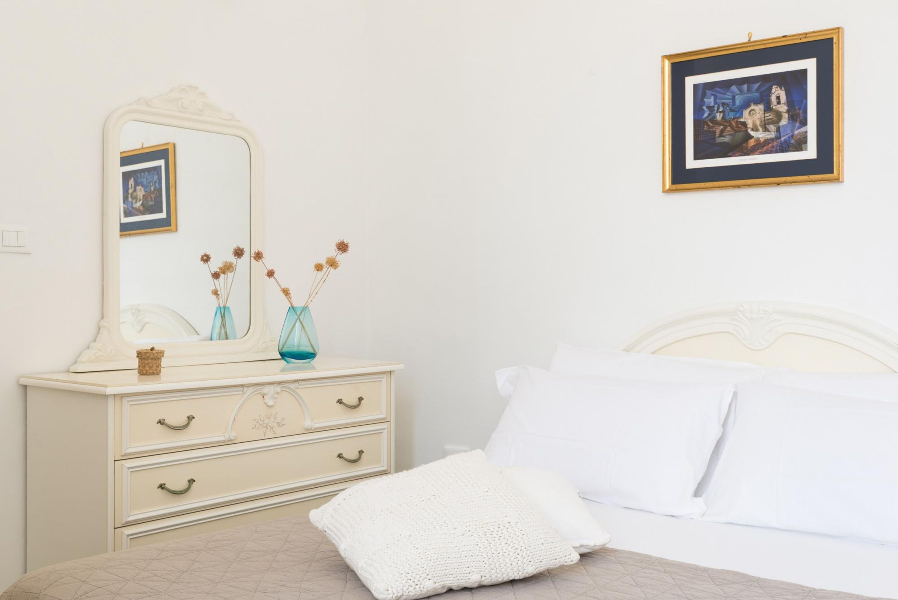 A bedroom with a white bed, a white dresser with drawers, a mirror, and a framed picture on the wall.