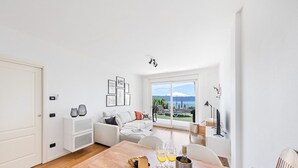 A bright, modern living room with a large window offering a scenic view, a cozy seating area, and a dining space with a wooden table. - Poggio al Sole 6 Apartment by Wonderful Italy (Toscolano Maderno)