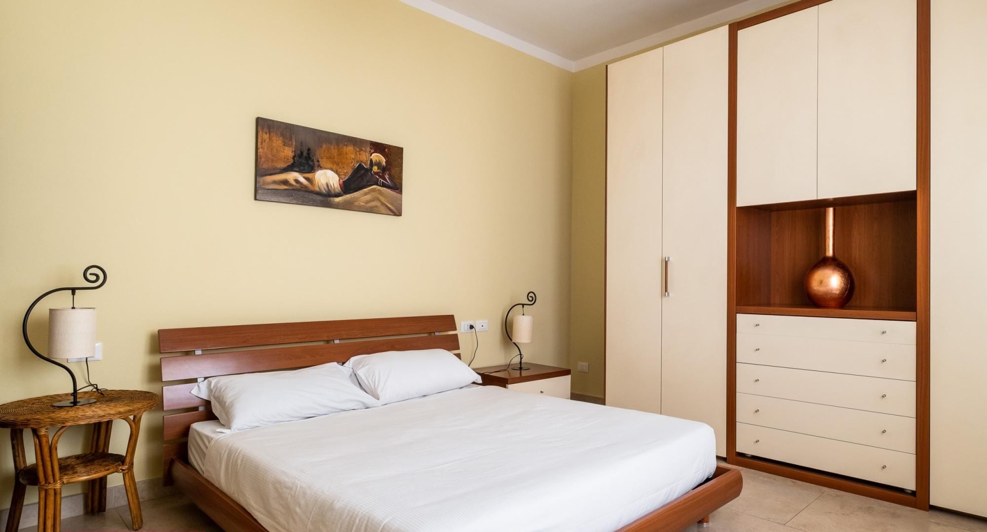 A bedroom with a bed, two bedside tables with lamps, a wardrobe, and a painting on the wall.