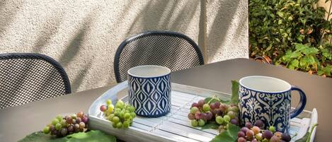 Una mesa con dos tazas decoradas, una bandeja con uvas y hojas, y sillas.