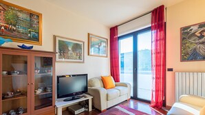 A hotel room with a television, a sofa, a cabinet with glass doors, and a large window with red curtains. - Kristall Lago 233 Apartment by Wonderful Italy (Desenzano del Garda)