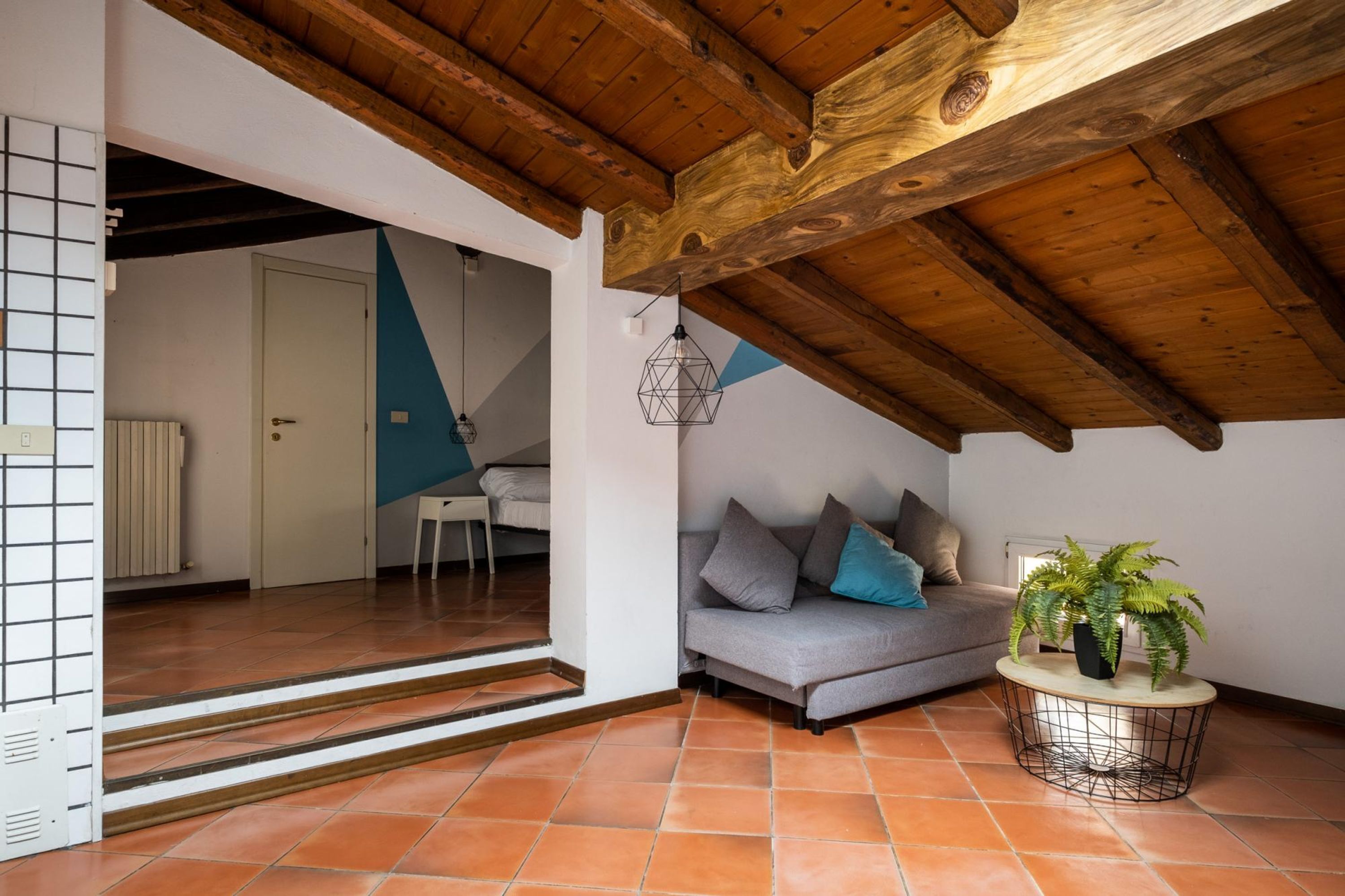 Een kamer met een houten plafond, een grijze bank met kussens, een klein rond tafeltje met een plant, en een witte deur die naar een andere kamer leidt.