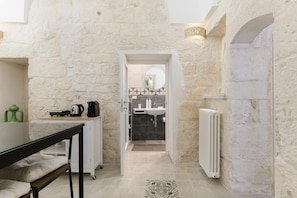 A stone-walled bathroom with a sink, mirror, and shower. A radiator is on the wall, and a table with a kettle and cups is visible in the foreground.