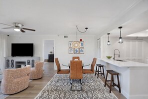 A modern living room with a flat-screen TV, patterned sofas, a dining area with a chandelier, and a kitchen with a bar.