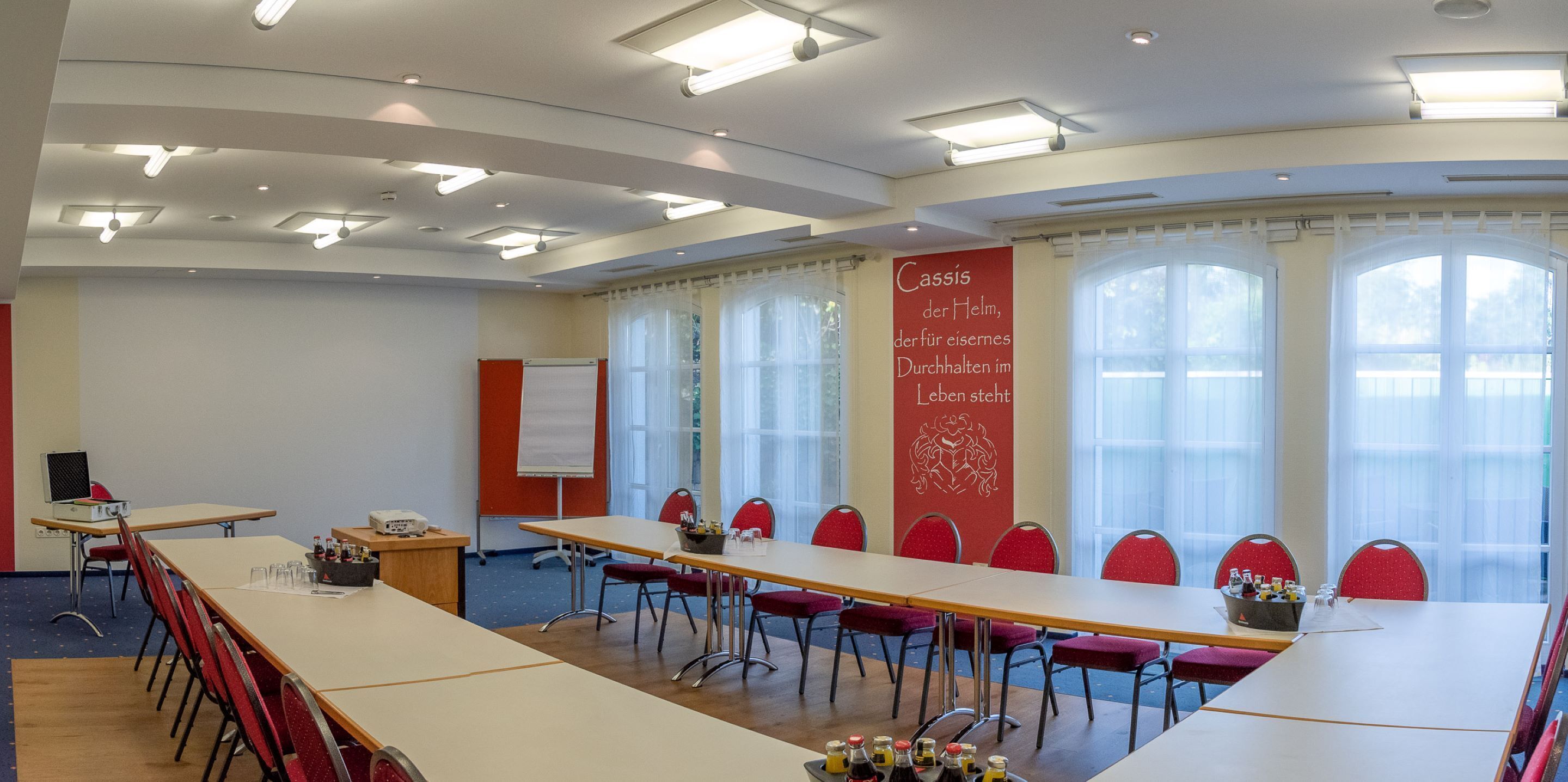 Un salón de conferencias con una larga mesa, sillas rojas, una pizarra y un cartel con texto.