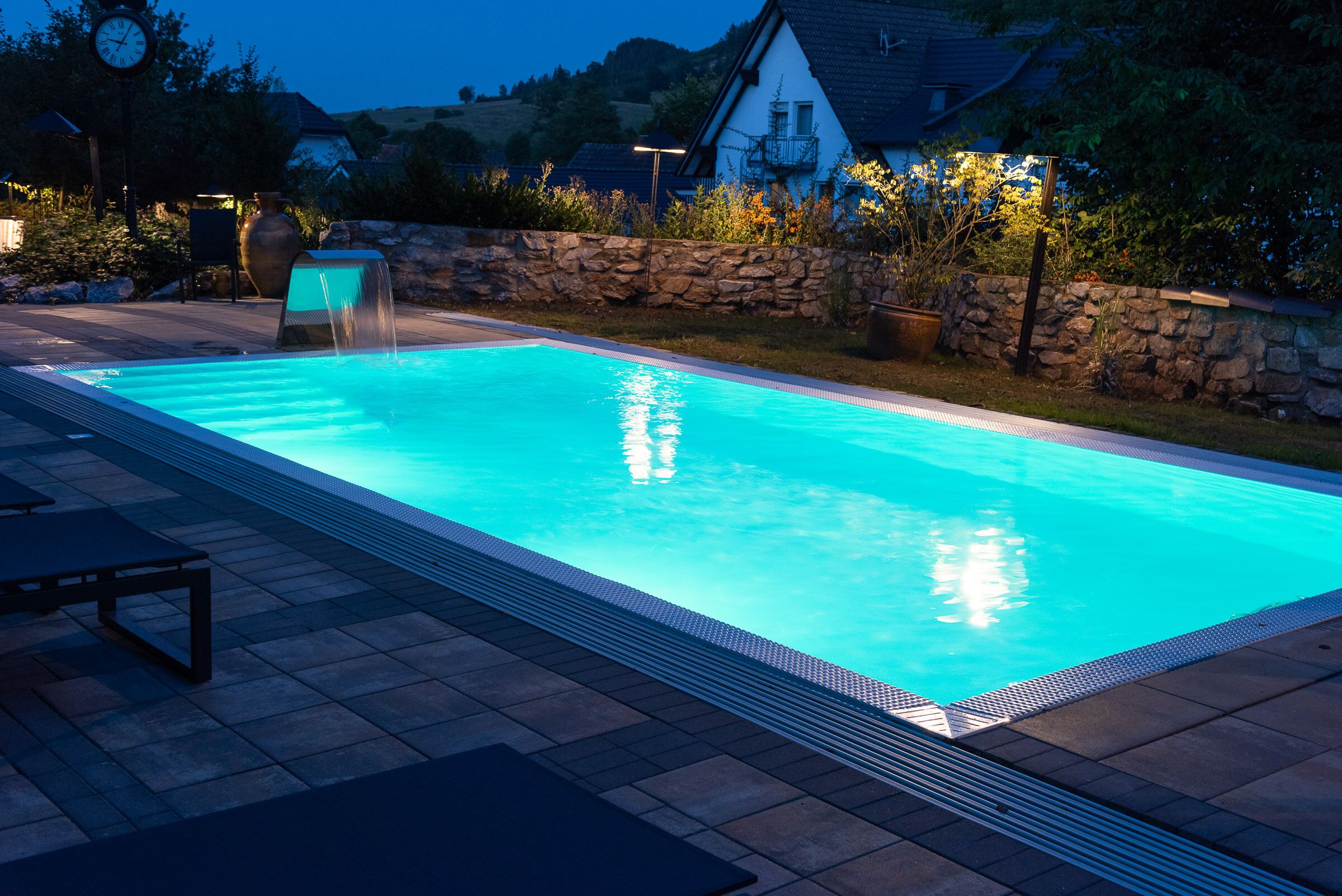 A rectangular swimming pool with a stone wall and a clock in the background.