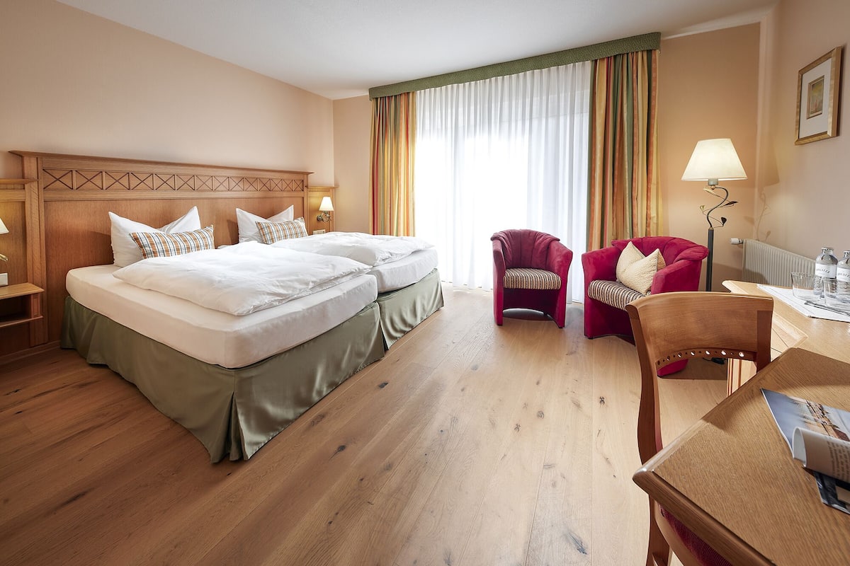 A hotel room with two beds, a desk, a chair, and a lamp.