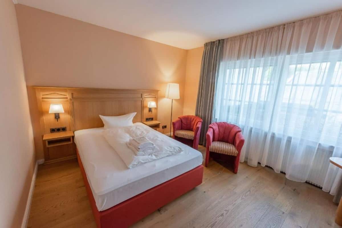 A hotel room with a large bed, two bedside lamps, a red chair, and a window with sheer curtains.