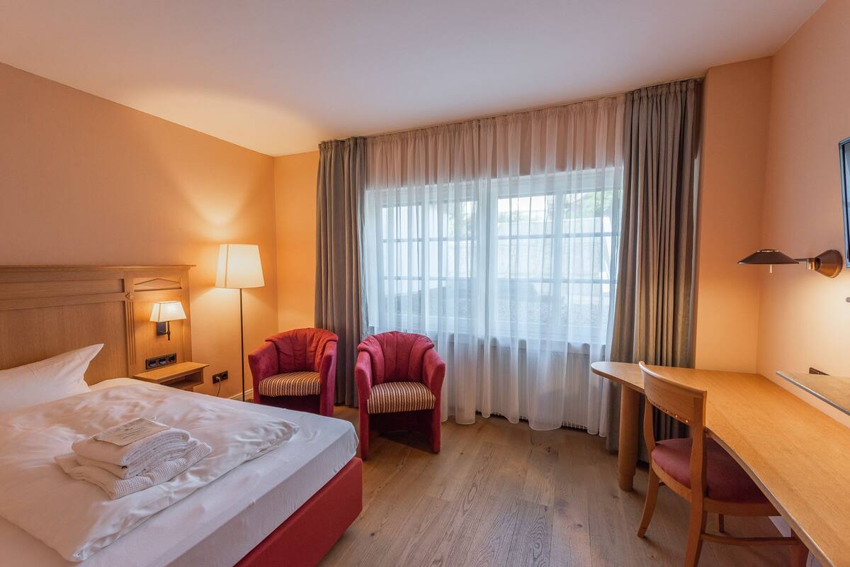 A hotel room with a bed, two armchairs, a desk, and a large window with curtains.