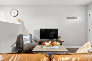 A living room with a sofa, a coffee table with flowers, a TV, a clock, and a coat rack. - Jan Dates Open: Pet friendly near 3 hospitals (Chattanooga)