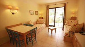 A cozy room with a dining table, chairs, a sofa, and a small table. There are two wall-mounted lamps, a sliding glass door leading to an outdoor area, and a small cabinet.