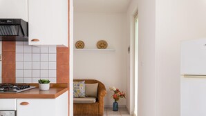 Una cucina con un fornello, un forno e una piccola pianta in vaso. C'è una sedia in vimini con un cuscino e un vaso con dei fiori. A parete, mensole con piatti decorativi.