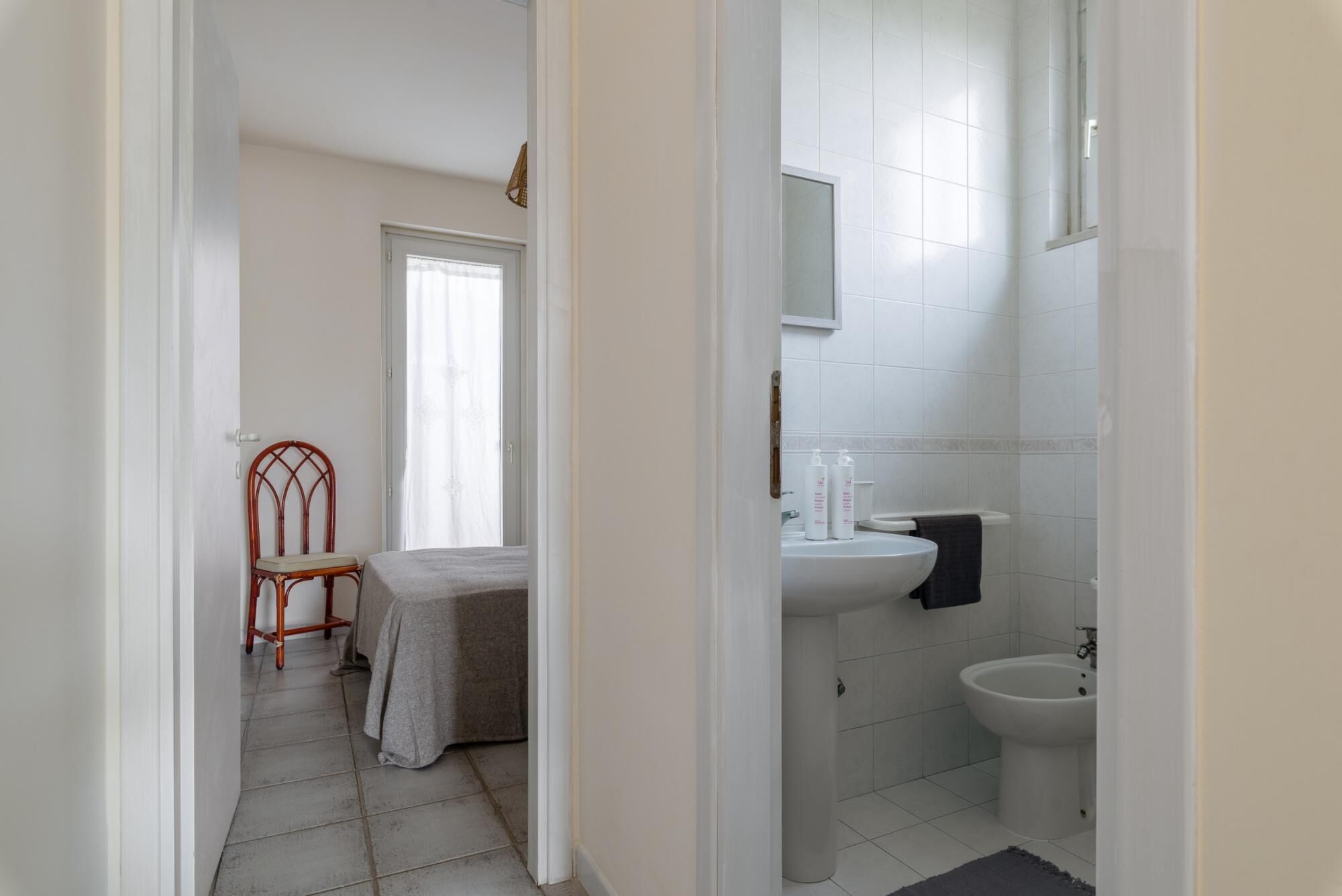 Een badkamer met een wastafel, bidet en spiegel. In de aangrenzende kamer staat een stoel en een tafel met een tafelkleed.