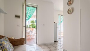 Una camera d'albergo con pavimento piastrellato, un divano con cuscini decorati, un cesto in vimini e una porta che dà su una zona esterna con sedie e un tavolo.