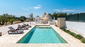 A swimming pool with lounge chairs, a white building with conical roofs, and a clear blue sky. - Trullo La Dolina con Piscina by Wonderful Italy (Ostuni)