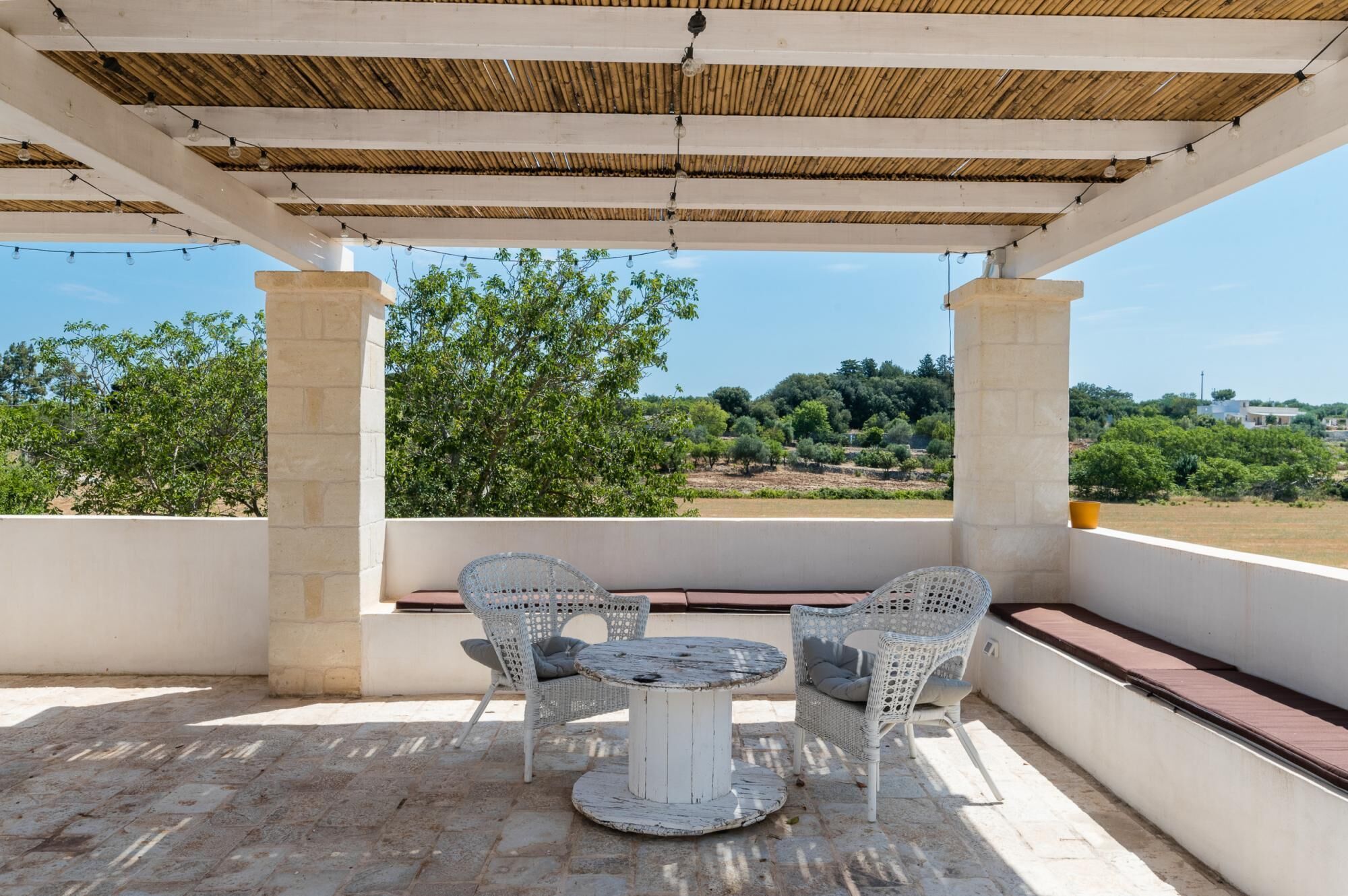 Un espacio al aire libre cubierto con una mesa redonda y sillas de mimbre, con vistas a un paisaje de árboles y edificios.