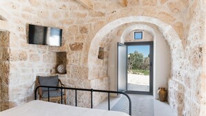 A stone-walled room with a bed, a television mounted on the wall, and a view of the outside through an open door. - Trullo La Dolina con Piscina by Wonderful Italy (Ostuni)