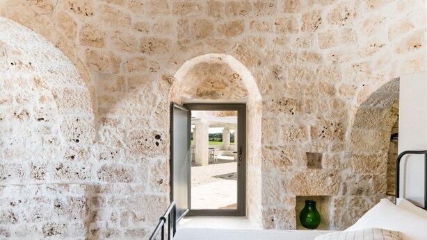 A stone-walled room with a bed, a green vase, and a view through an archway to an outdoor area. - Trullo La Dolina con Piscina by Wonderful Italy (Ostuni)