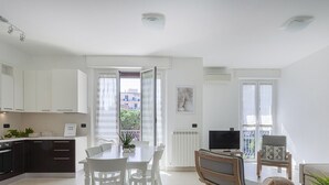 Un appartement moderne comprenant une cuisine, un coin repas et un salon. Il dispose d’un balcon offrant une vue sur les immeubles et les arbres.