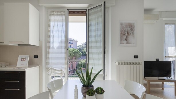 Une cuisine moderne avec des meubles blancs, une table à manger avec des chaises, une plante en pot et un téléviseur en arrière-plan.