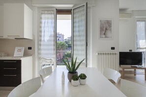 Uma cozinha moderna com armários brancos, uma mesa de jantar com cadeiras, uma planta em vaso e uma televisão ao fundo.