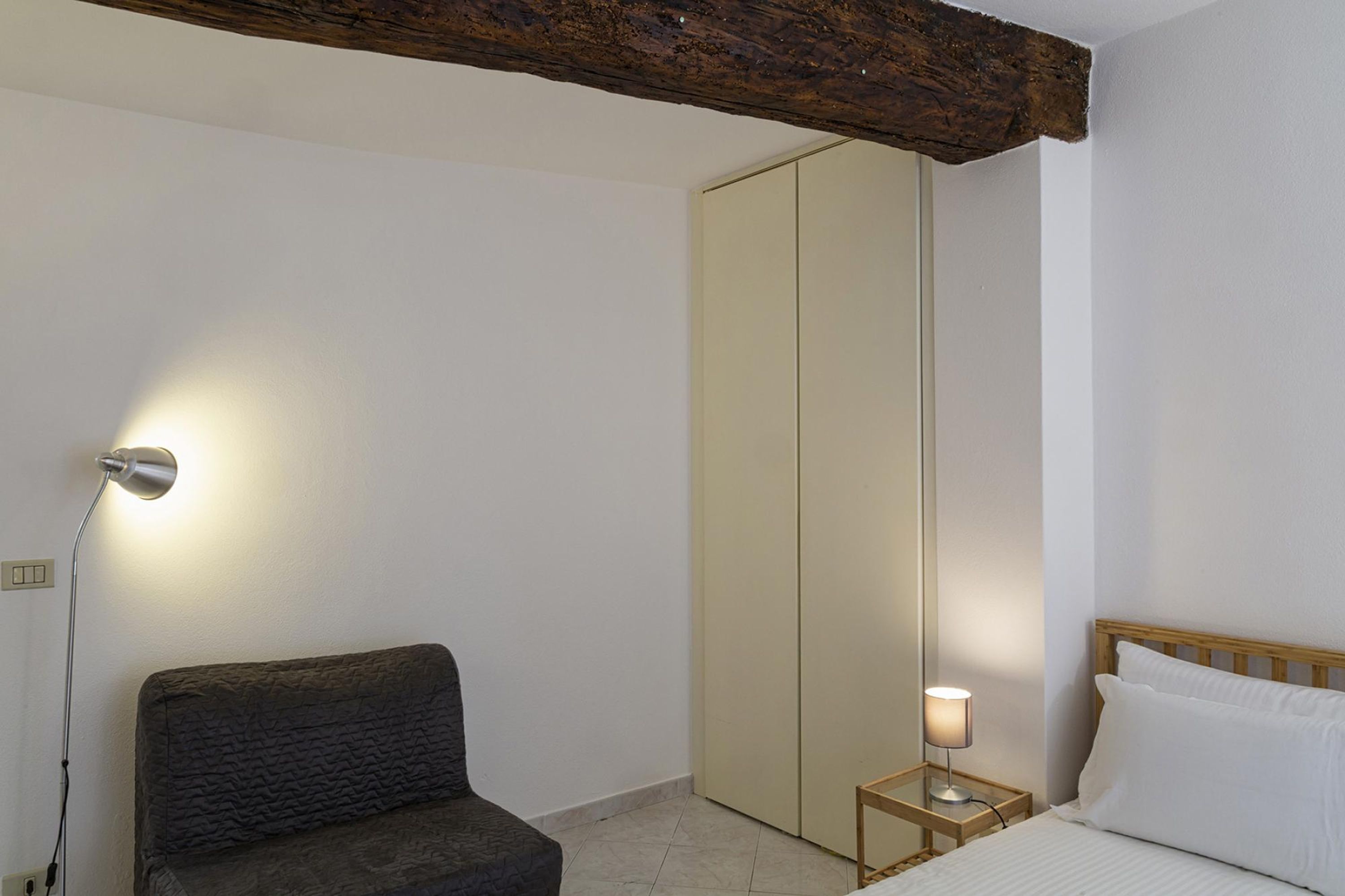 A bedroom with a bed, a bedside table with a lamp, a dark sofa, and a wardrobe.
