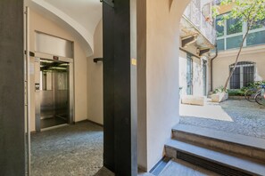 Un pasillo interior con un ascensor y una escalera que lleva a un área exterior con una bicicleta y plantas en macetas.