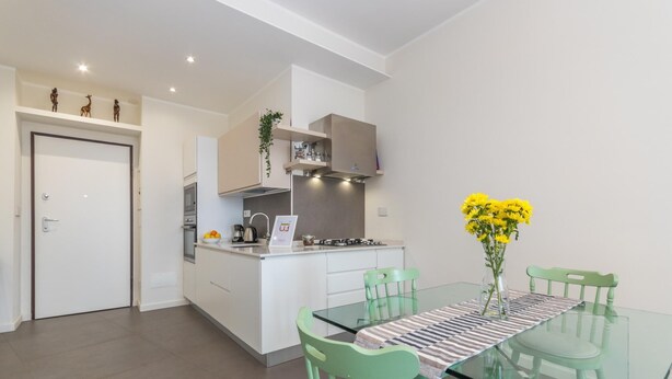 A modern kitchen with white cabinets, a built-in oven, and a gas stove. There is a dining area with a glass table and green chairs.