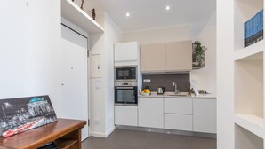 A modern kitchen with white cabinets, a built-in oven, and a microwave. There is a small countertop with a kettle and a bowl of fruit.