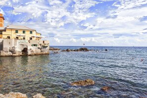 A coastal building with a clear view of the sea, a flag on a rock, and a distant ship.