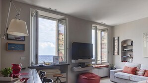 Una sala de estar moderna con ventanas amplias, un televisor de pantalla plana, una zona de comedor con una jarra roja y una acogedora sala de estar con un sofá blanco y cojines rojos.