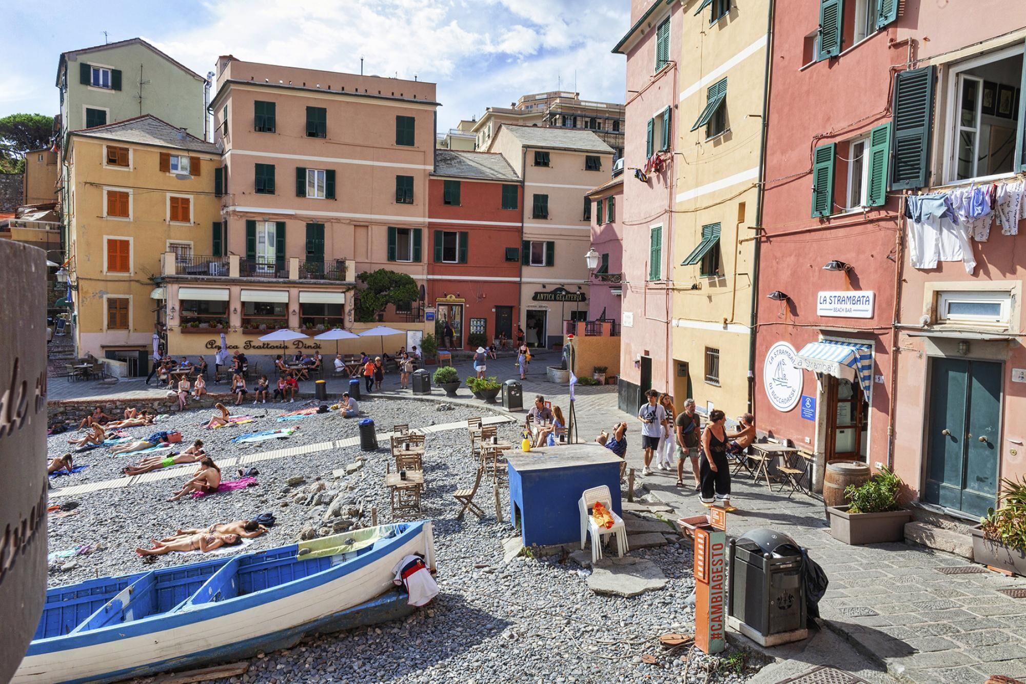 A beachside piazza with people sunbathing, a small boat, and buildings with shops and cafes.