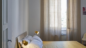 Een slaapkamer met een bed, een bedlampje, een raam met gordijnen, en een stoel.