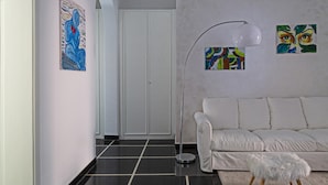 Een moderne woonkamer met een witte bank, een zachte witte poef en een vloerlamp. Aan de muren hangen abstracte schilderijen.
