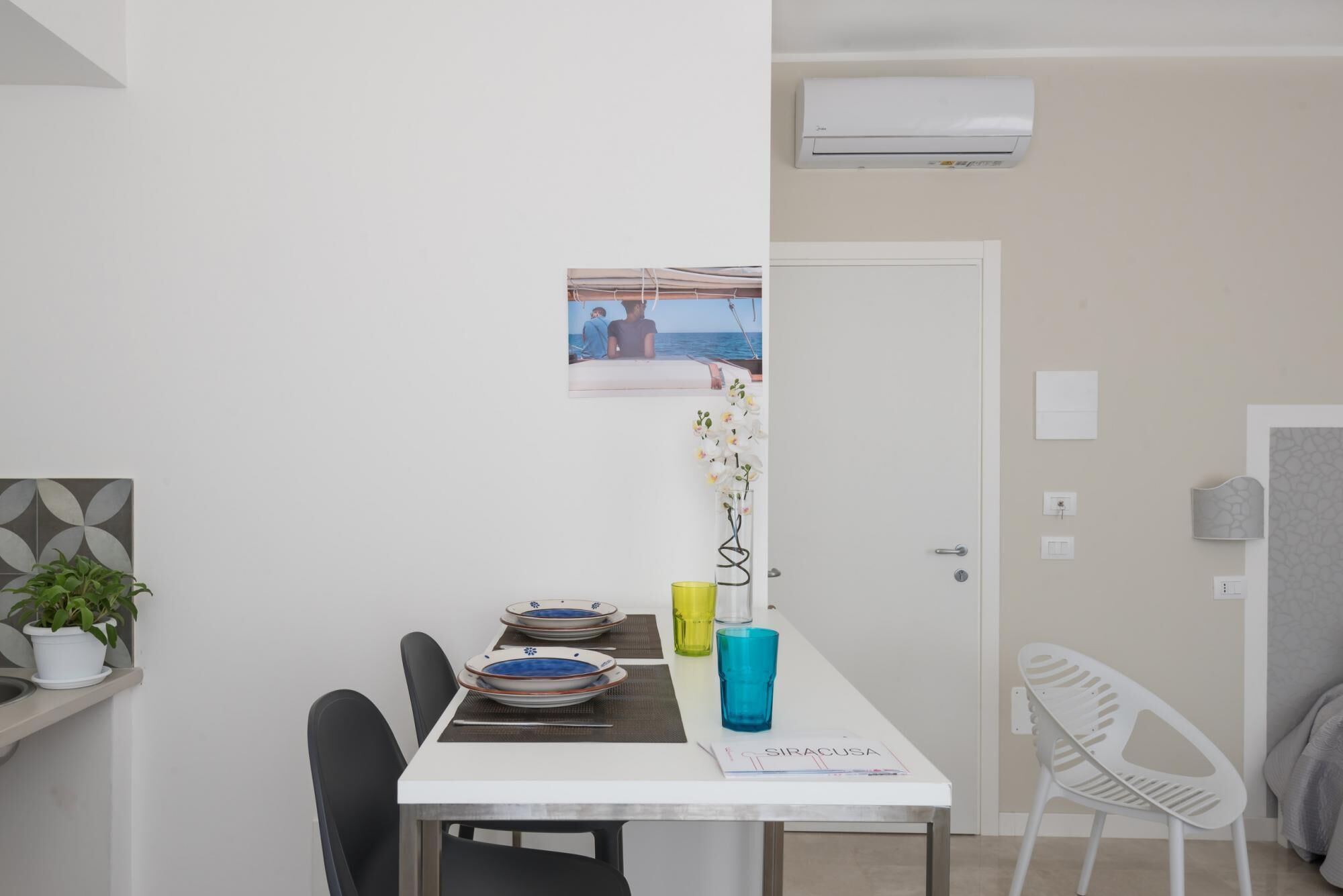 Un coin repas avec une table blanche, des chaises noires, un tableau au mur et une plante en pot.