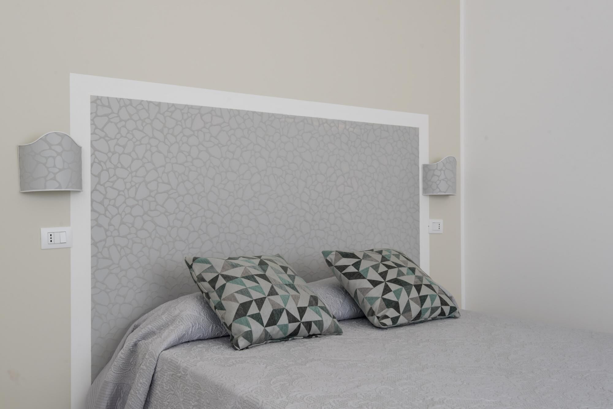 Ein Schlafzimmer mit einem Bett, zwei gemusterten Kissen, einem strukturierten Kopfteil und Wandleuchten.