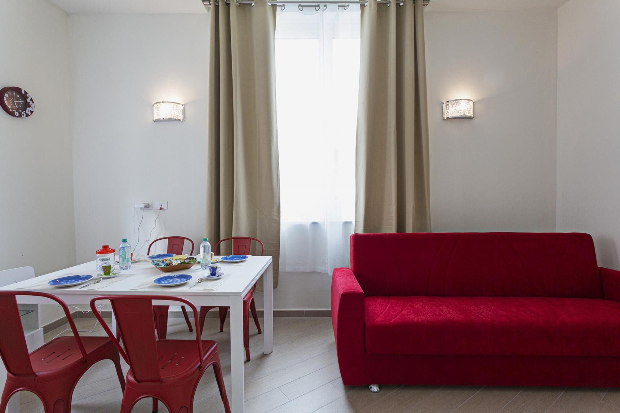 A dining area with a white table, red chairs, a red sofa, and beige curtains.