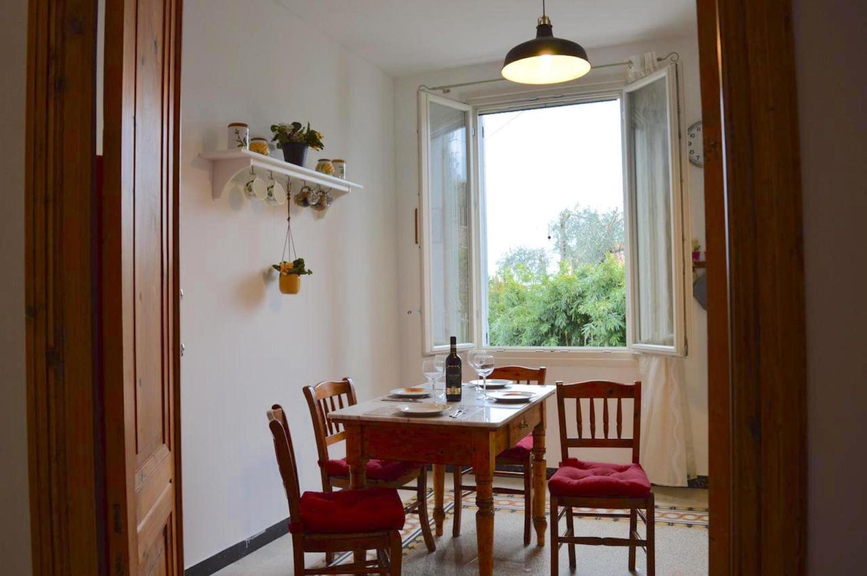 Een eethoek met een tafel gedekt voor twee personen, een raam met uitzicht op groen, en een plank met decoratieve spullen.