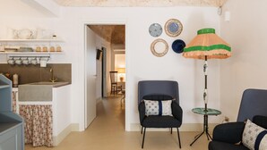 Een moderne woonkamer met een donkerblauwe fauteuil, een ronde tafel en een vloerlamp met een gestreept lantaarn. In de achtergrond is een keuken met een spoelbak en planken zichtbaar.