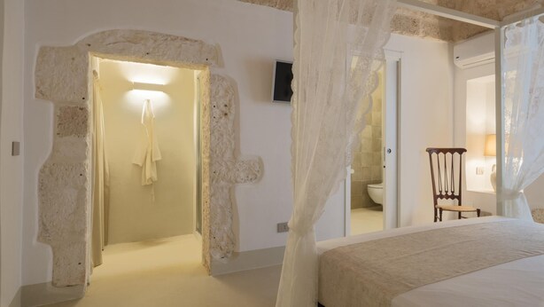 Een slaapkamer met een bed, een stoel, een spiegel en een badkamer zichtbaar door een boog.