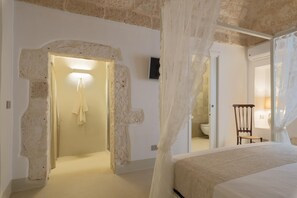 A bedroom with a bed, a chair, a mirror, and a bathroom visible through an archway. - Cementine Traditional Suites - 9 - CAV (Ostuni)