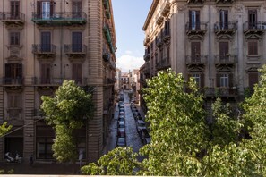 A narrow street flanked by multi-story buildings with balconies, parked cars, and trees.