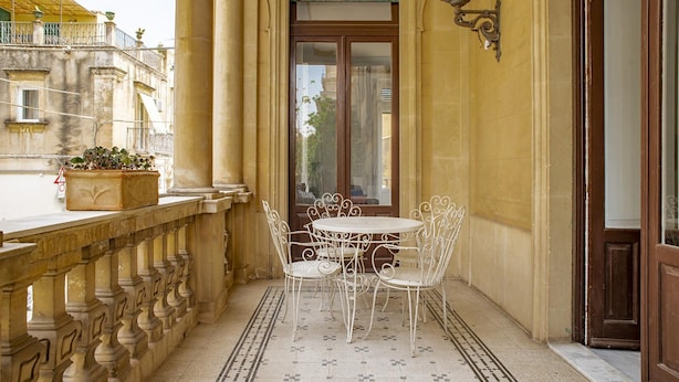 Uma varanda com uma mesa e cadeiras de metal branco, um vaso de planta e vista para a rua.