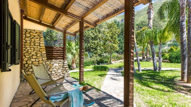 A covered patio with stone walls, a stone floor, and a view of a lush garden with palm trees. - L Ulivo Blu - One-bedroom With Garden by Wonderful Italy (Castellammare del Golfo)
