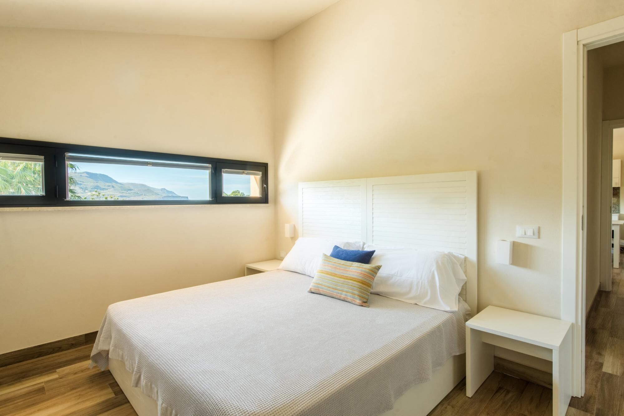 A bedroom with a bed, a nightstand, a window with a view, and a door to another room.