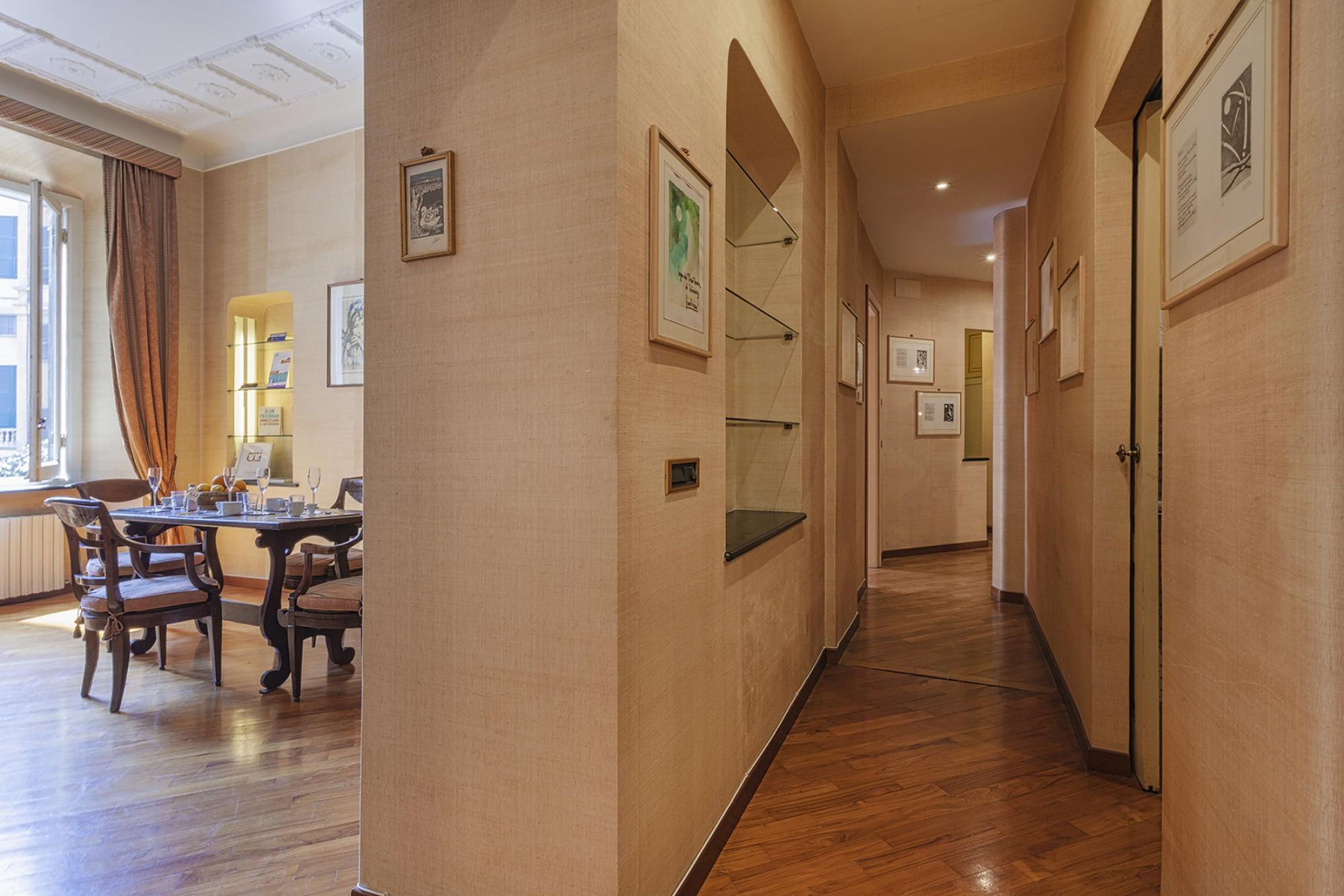 A hallway with wooden flooring, framed artwork on the walls, and a dining area with a table and chairs.