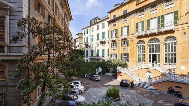 Un cortile con auto e motociclette parcheggiate, circondato da edifici con persiane verdi e una scala centrale.