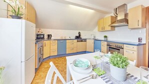 Een moderne keuken met lichte houten kastjes, een blauw aanrecht en een eethoek met een witte tafel en stoelen.